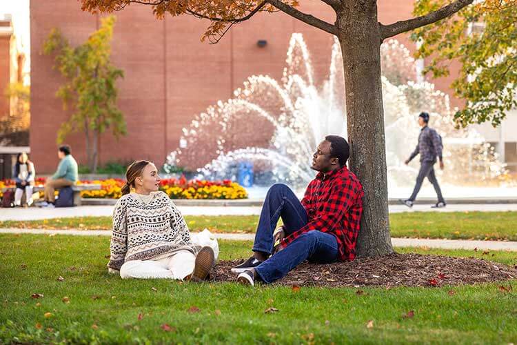 Two students sitting under a tree near the UWM Fountain on a nice Fall day talking to each other.